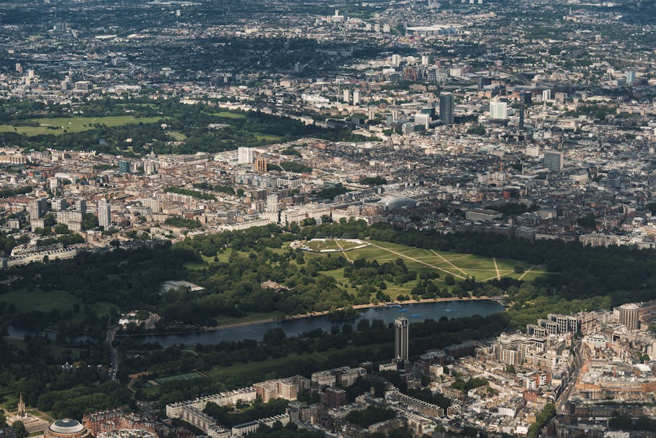 An aerial view of central London showing the expansive green space of Hyde Park with its walkways, surrounded by dense urban development including residential and commercial buildings. A river runs al