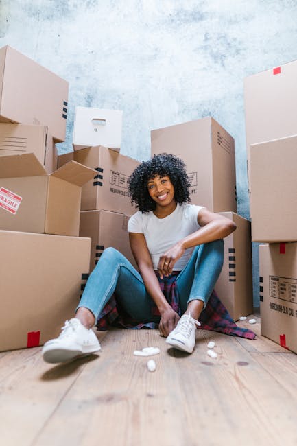 A man with curly hair and a beard, wearing a blue t-shirt and jeans, is sitting on the wooden floor of a room next to a stack of cardboard moving boxes, some sealed with red tape. The room has large,